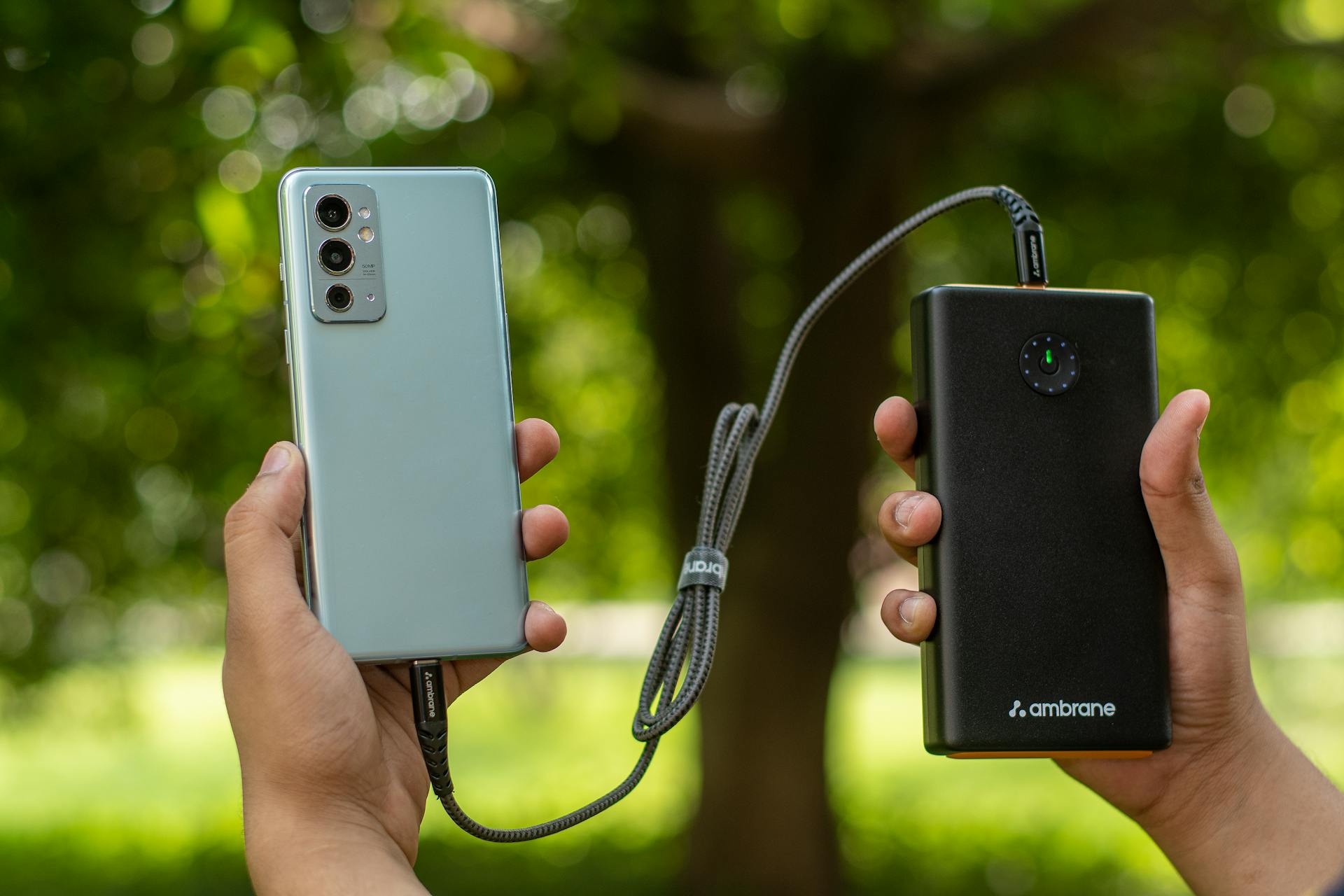 A person holds a smartphone in one hand and a black power bank in the other, both connected by a charging cable, against a green backdrop.