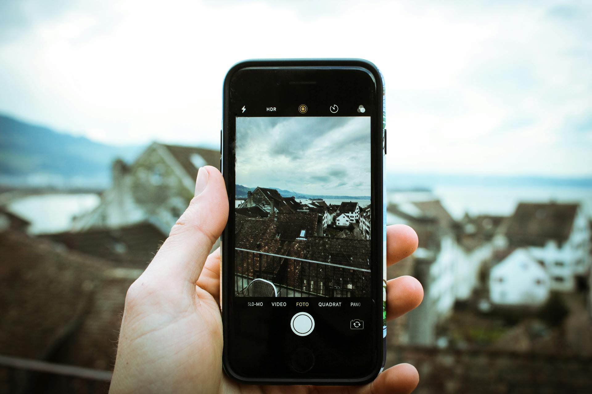 A hand holding a smartphone displays a scenic view of rooftops and a lake under a cloudy sky.