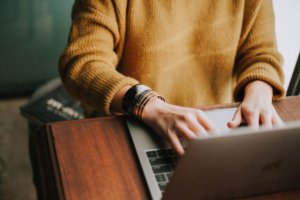 A person in a mustard sweater types on a laptop, adorned with gold and black bracelets, with books visible in the background.
