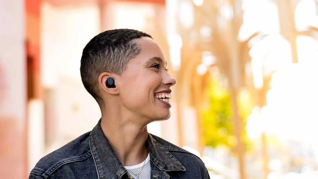 A young person wearing stylish wireless earbuds, standing outdoors with a softly blurred background of warm colors.