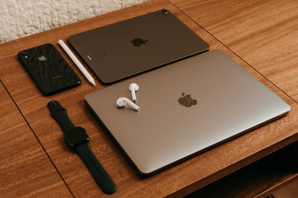 A flat lay of Apple devices: an iPhone, iPad with Apple Pencil, MacBook, AirPods, and an Apple Watch, all on a wooden table.