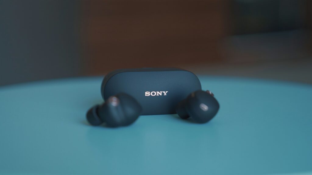 A pair of Sony wireless earbuds rests beside their charging case on a turquoise surface, highlighted against a blurred background.