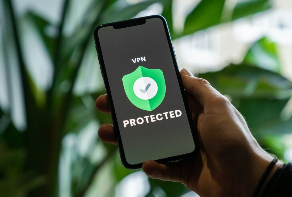 A hand holding a smartphone displaying a 'VPN Protected' logo with a green shield and a checkmark, set against green foliage.