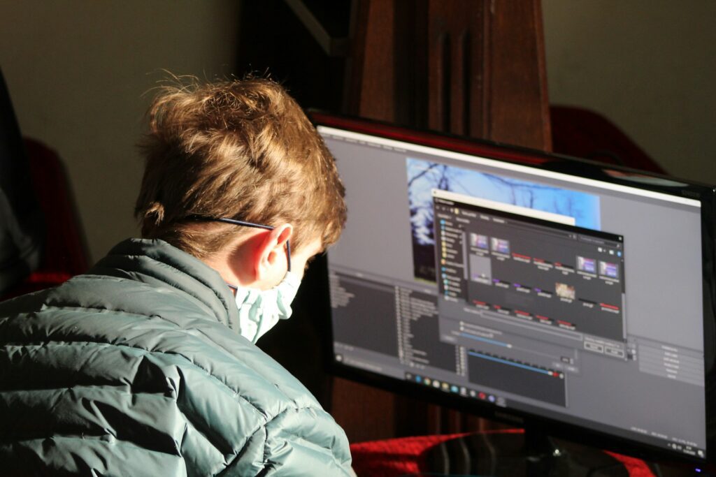 A person wearing a jacket and a mask is focused on a computer screen displaying editing software and media files.