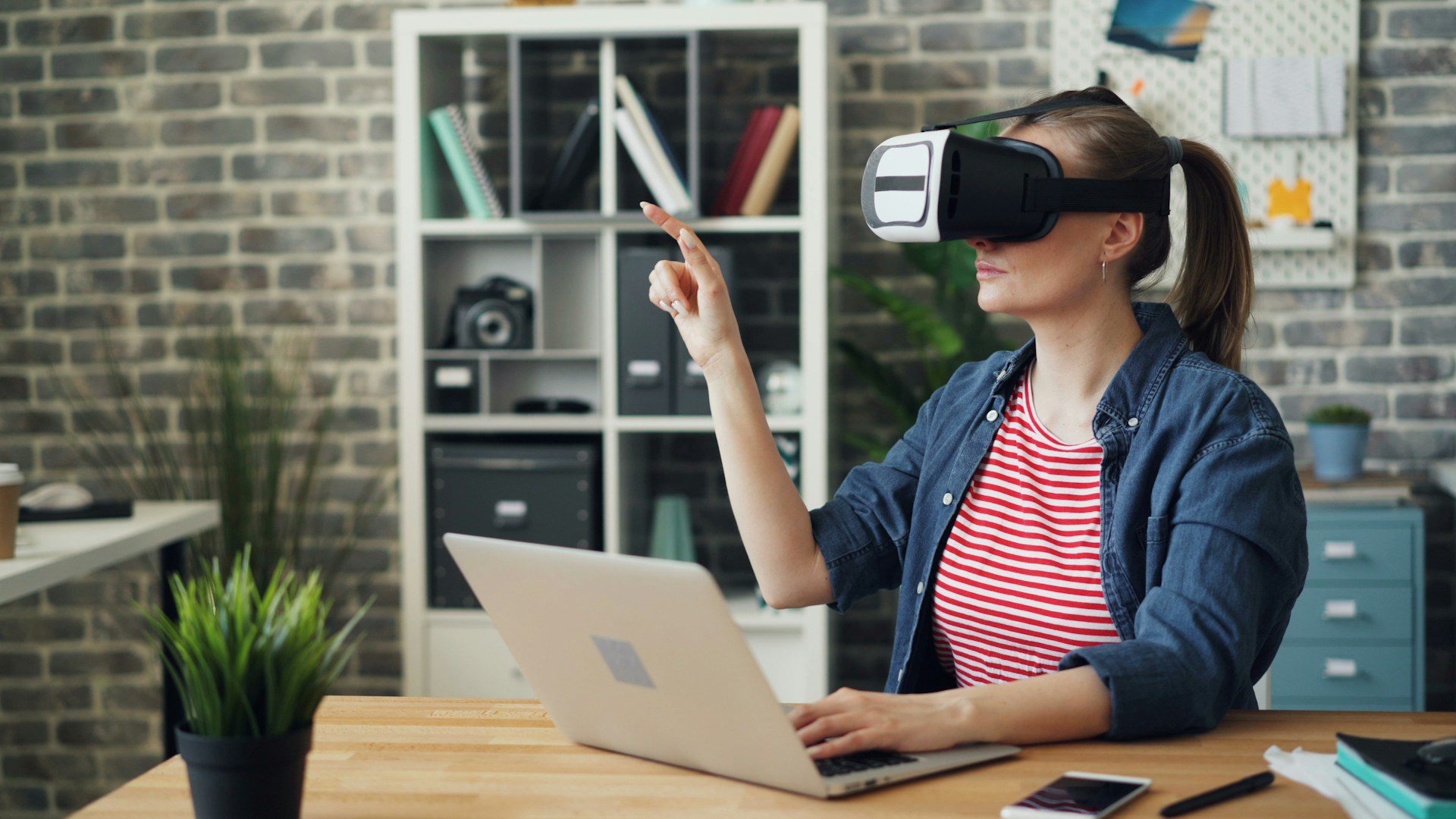 A person wearing a VR headset interacts with a laptop in a modern workspace surrounded by plants and shelves with various items.