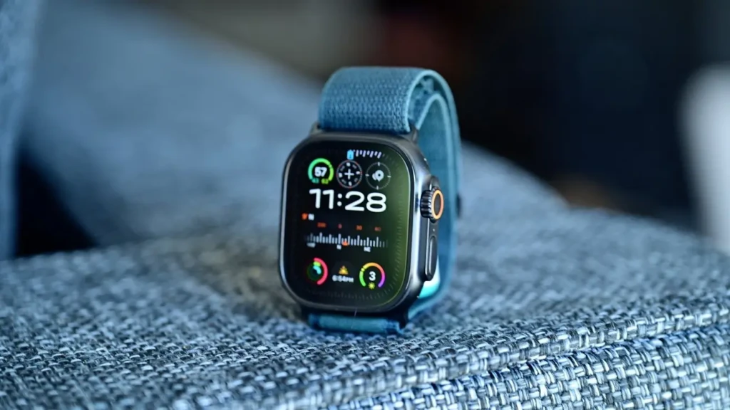 An Apple Watch Ultra with a blue woven band rests on a textured blue fabric surface. The screen shows a detailed watch face with the time (11:28), activity rings, and various complications for weather and fitness tracking.