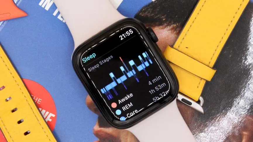 A close-up of an Apple Watch with a light pink band, resting on a colorful magazine cover next to a yellow strap. The watch face displays the Sleep app, showing a detailed graph of sleep stages (Awake, REM, Core) for the night.