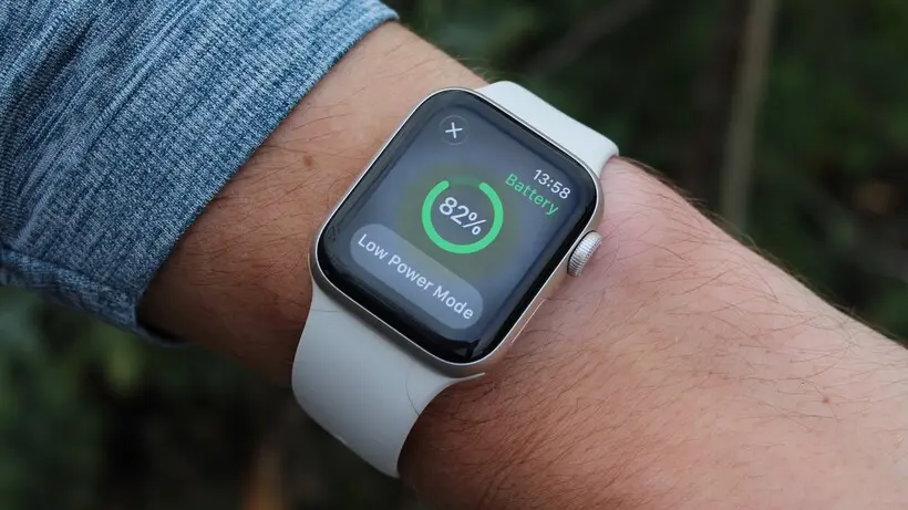An Apple Watch with a white band is shown on a person's wrist, displaying the Battery app. The screen shows an 82% charge and indicates that Low Power Mode is active, with the time reading 13:58.