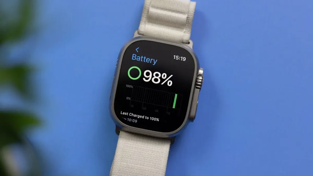 An Apple Watch Ultra with a light gray band is shown against a solid blue background. The screen displays the Battery app, indicating a 98% charge and showing a graph of recent battery usage, with the time reading 15:19.
