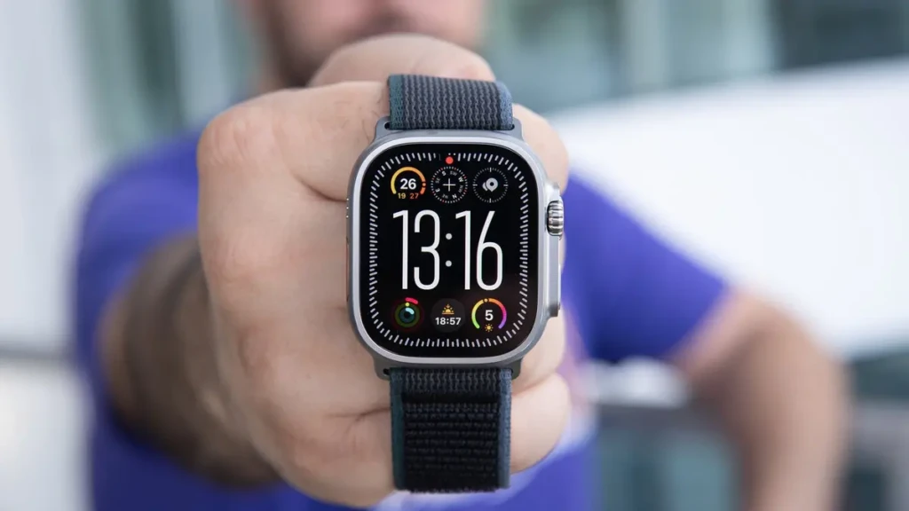 A person holds an Apple Watch Ultra with a dark gray band directly towards the camera. The watch face displays a bold digital time (13:16) surrounded by multiple activity and weather complications, set against a blurred outdoor background.