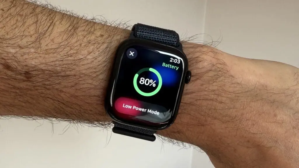 An Apple Watch with a black band is shown on a person's wrist, displaying the Battery app. The screen shows an 80% charge and a red "Low Power Mode" button, with the time reading 2:03.