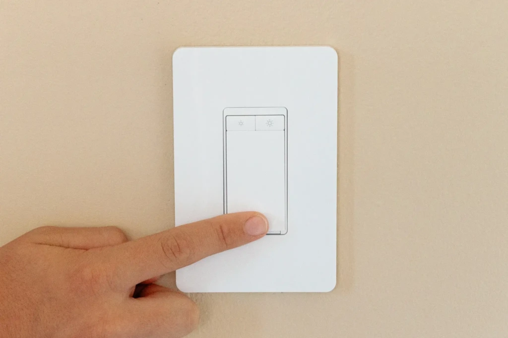 A hand pressing a modern light switch on a beige wall, featuring icons for brightness control.