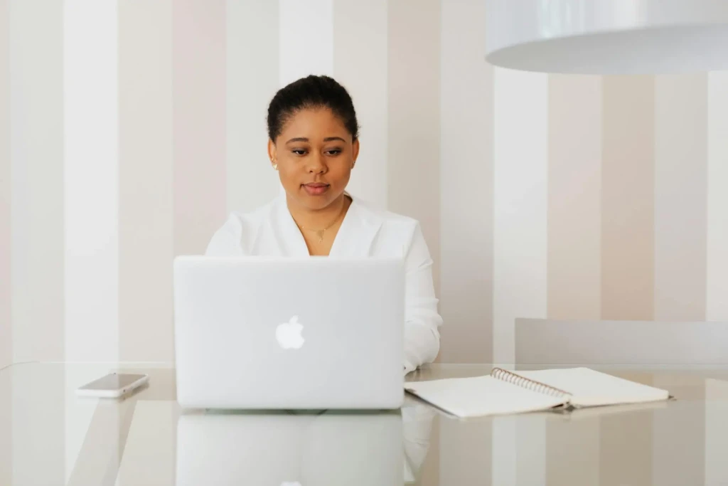 A professional individual in a white blouse sits at a glass table, working on a laptop, with a notebook and smartphone nearby.