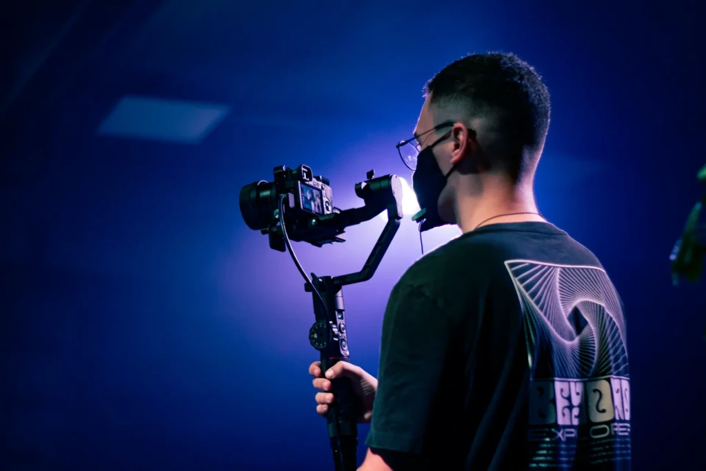A person holds a camera on a stabilizer, set against an abstract blue-lit background, wearing a mask and glasses.