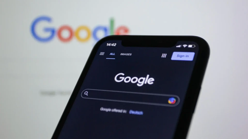 A close-up of a smartphone displaying the Google search homepage in dark mode. The Google logo is visible on the screen, and a larger, blurred Google logo appears in the background, suggesting a focus on the company's digital services.