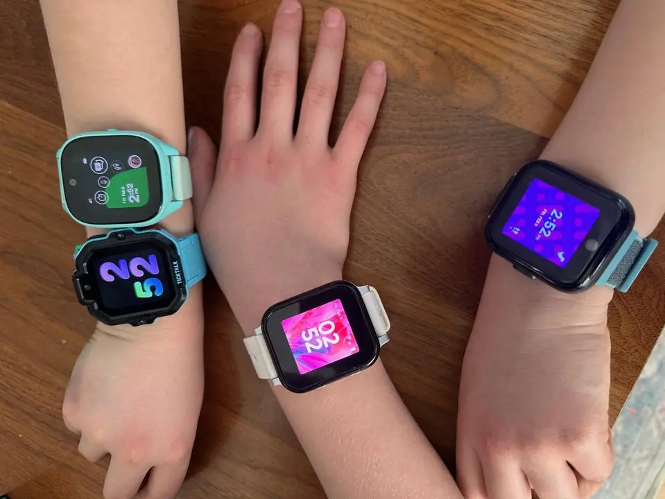 Three children's smartwatches are shown on different wrists, displaying colorful digital screens with various numbers and designs.