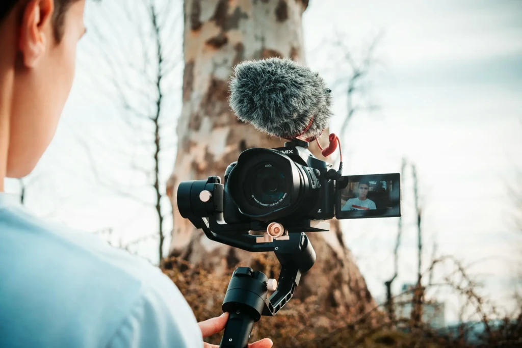 A person holds a camera mounted on a stabilizer, featuring a microphone, pointed toward a scenic background of trees.
