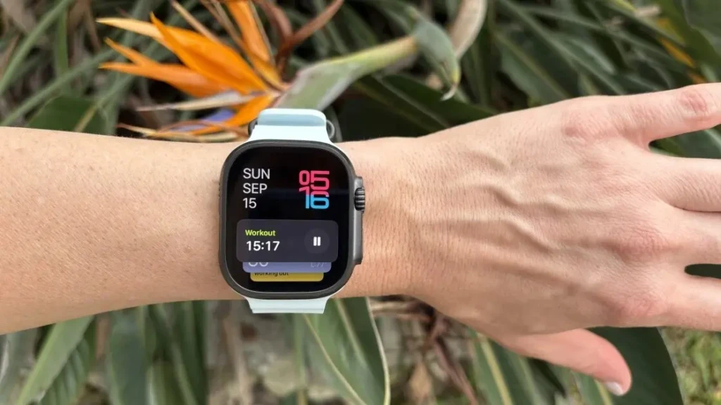 An Apple Watch Ultra with a light blue band is worn on a person's wrist outdoors, with vibrant orange flowers in the background. The screen displays a workout timer at 15:17, along with the date "SUN SEP 15" and a large digital clock.