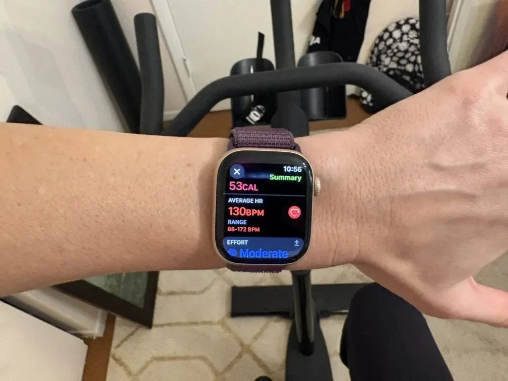 An Apple Watch with a purple woven band is worn on a person's wrist during a workout. The screen displays a summary of activity metrics, including 53 calories burned, an average heart rate of 130 BPM, and a moderate effort level, with the time at 10:56.