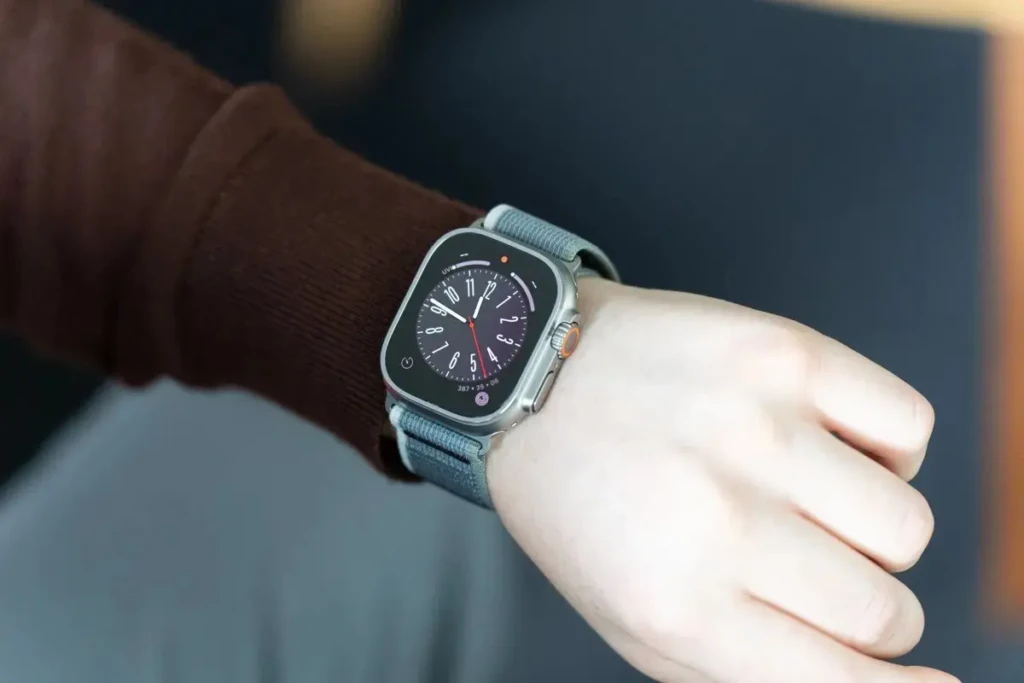 A close-up shot of an Apple Watch Ultra on a person's wrist, who is wearing a brown sweater. The watch features a classic analog face with white numerals and hands, paired with a gray sport loop band.