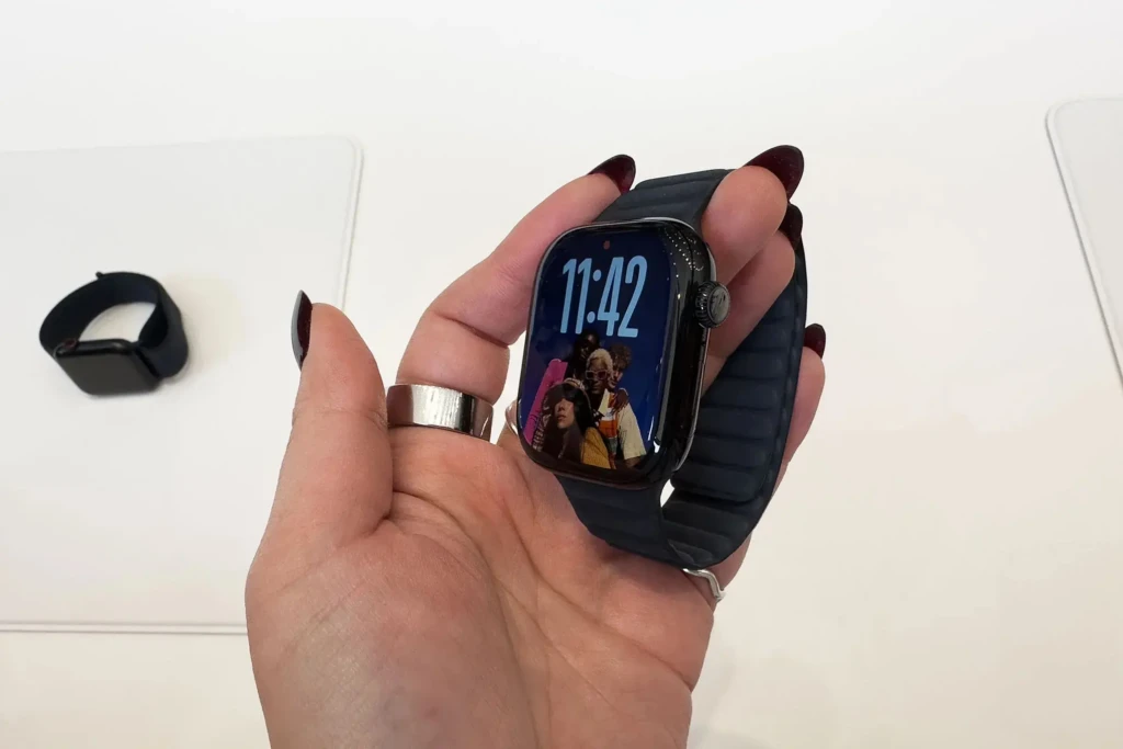 A hand with dark red nail polish holds an Apple Watch with a black case and band. The screen displays a photo-based watch face showing a group of people, with the time reading 11:42. Another watch is visible in the background on a white surface.