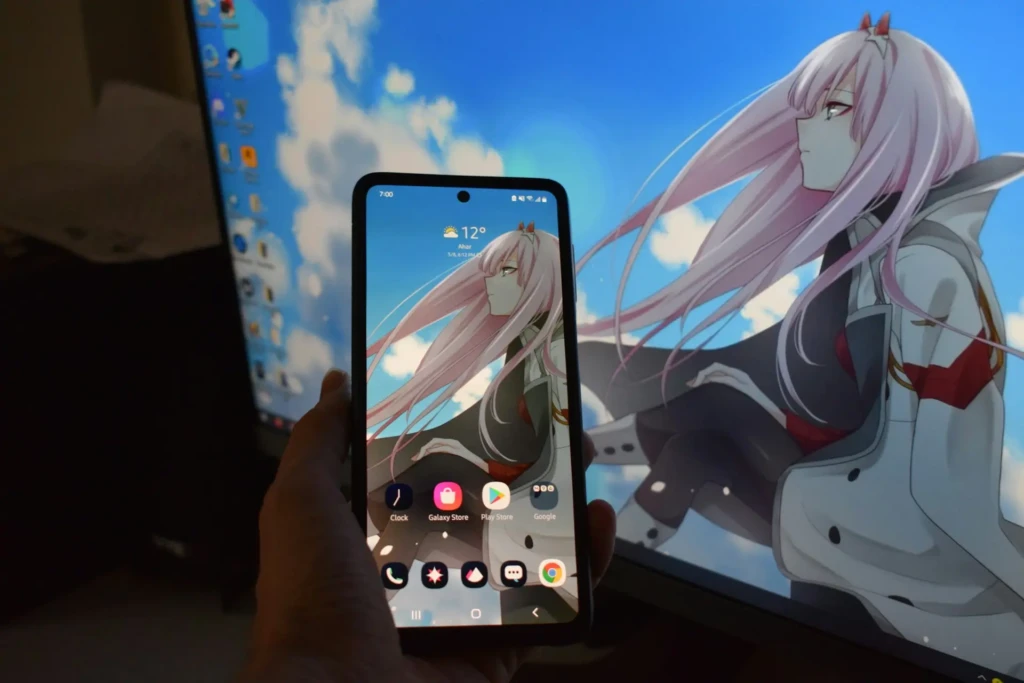 A hand holds an Android smartphone with a custom anime wallpaper featuring a pink-haired character. The phone's home screen shows app icons like Galaxy Store and Play Store. In the background, a computer monitor displays the same anime wallpaper, creating a matching aesthetic.