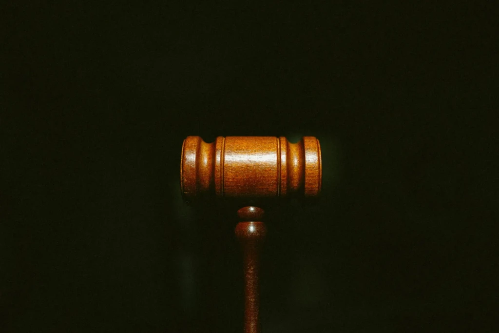 A close-up shot of a polished wooden judge's gavel against a solid black background. The lighting highlights the texture and grain of the wood, emphasizing its role as a symbol of legal authority and judgment.