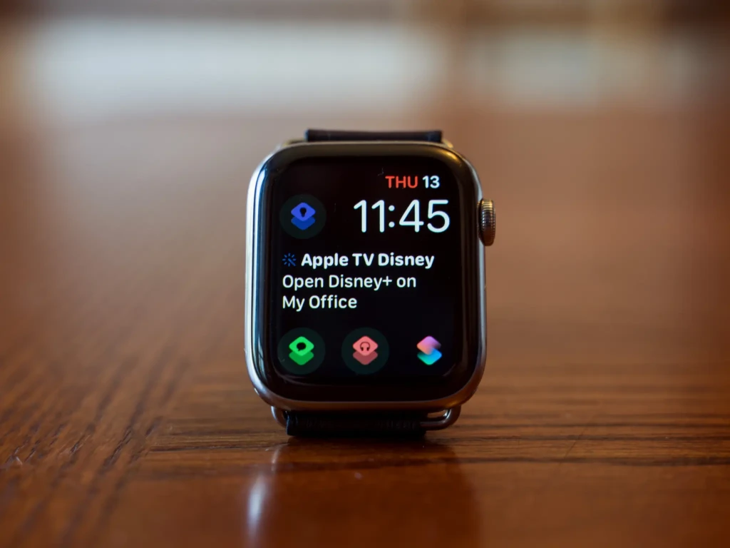 An Apple Watch with a brown leather band stands upright on a polished wooden surface. Its screen displays the time as 11:45 on Thursday the 13th, along with a notification to "Open Disney+ on My Office" and three app icons at the bottom.