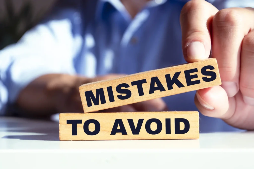 A hand holds a wooden block labeled "MISTAKES" above another block labeled "TO AVOID," with a blurred background of a shirt and table.