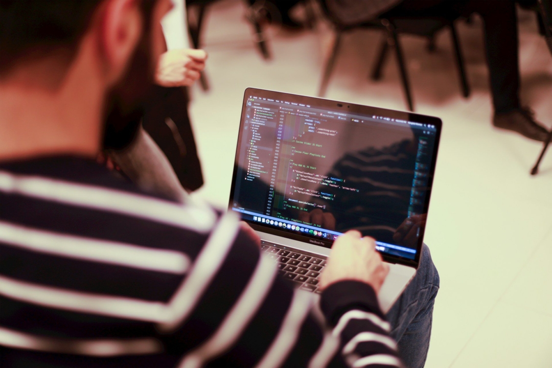 A person in a striped sweater coding on a MacBook Pro, with visible lines of code on the screen, in a group setting.