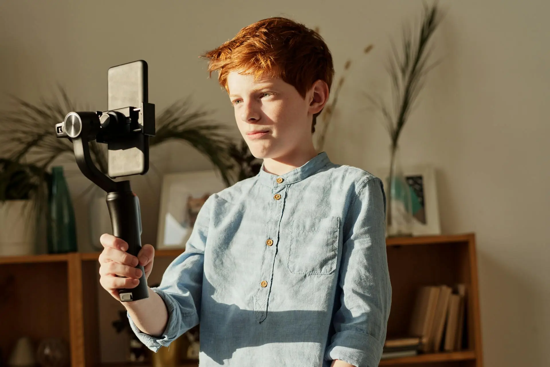 A child stands indoors, holding a smartphone stabilizer, wearing a light blue button-up shirt, with plants and shelves in the background.