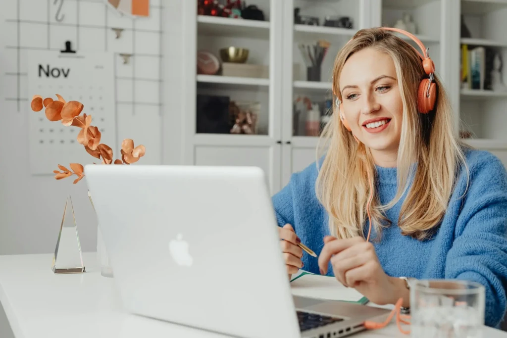 A person in a blue sweater wears headphones while working at a laptop, surrounded by plants and decorative items in a bright room.