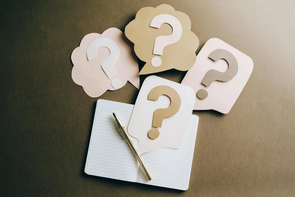 Four question mark speech bubbles in pink and beige, placed around a blank notebook and a gold pen on a brown surface.