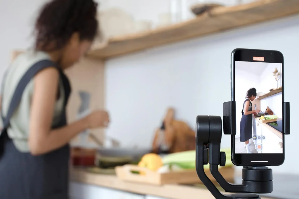 A smartphone mounted on a gimbal records a person cooking in a modern kitchen, surrounded by fresh ingredients.
