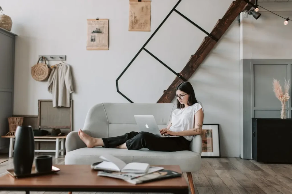 A woman with glasses and a tattoo on her arm is sitting comfortably on a gray sofa, using a silver MacBook. The setting is a stylish, modern living room with wooden floors, a staircase, and decorative items like framed art and a woven bag.