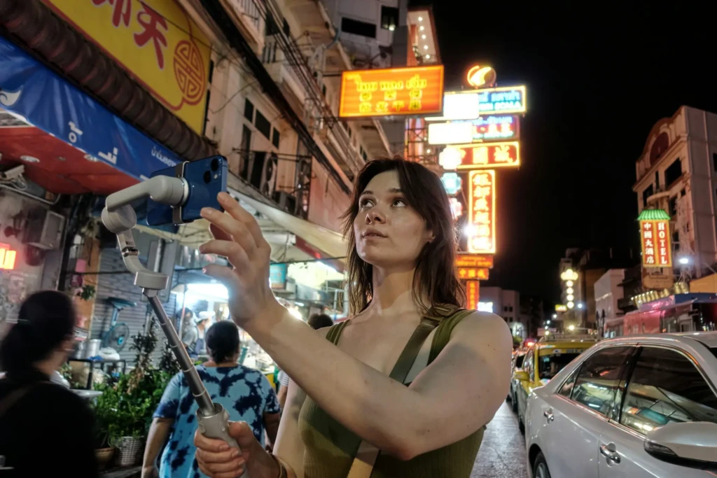 A person holds a smartphone on a selfie stick in a vibrant, bustling night market, illuminated by colorful neon signs.