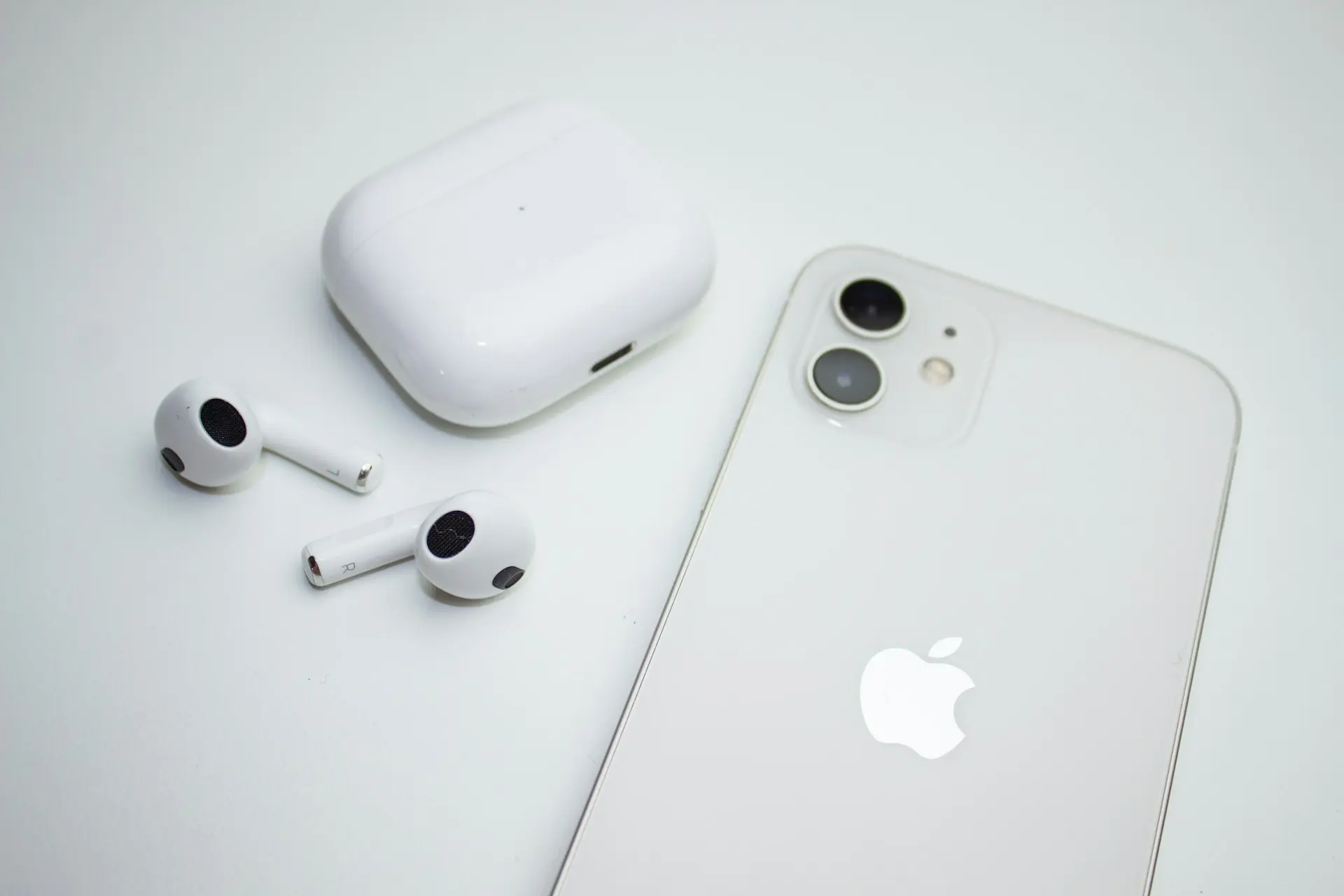 A top-down view of a white iPhone placed next to a pair of white AirPods and their charging case on a clean white surface. The Apple logo is visible on the back of the phone.