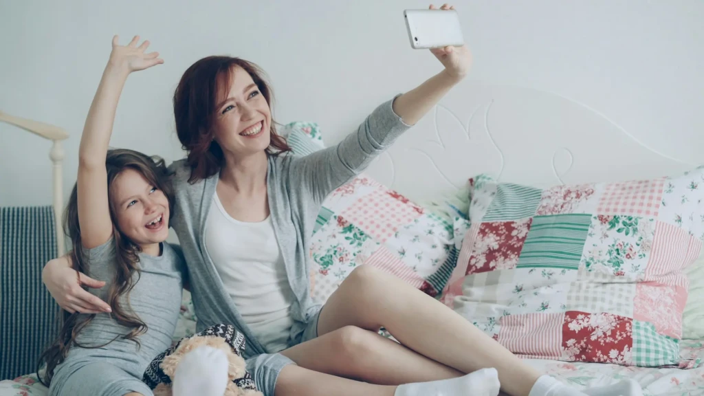 A woman and a girl take a selfie together on a cozy bed adorned with colorful patchwork pillows. Both appear joyful and relaxed.
