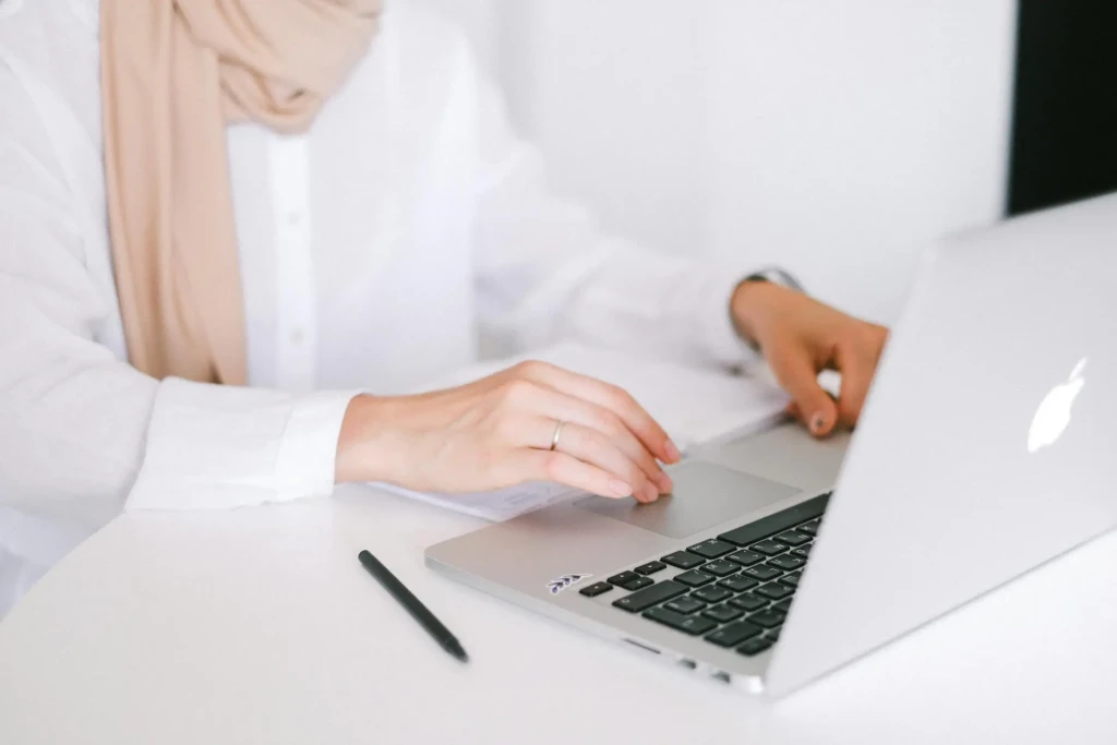 A person in a white shirt and scarf uses a laptop at a desk, with a pen nearby and papers in hand.