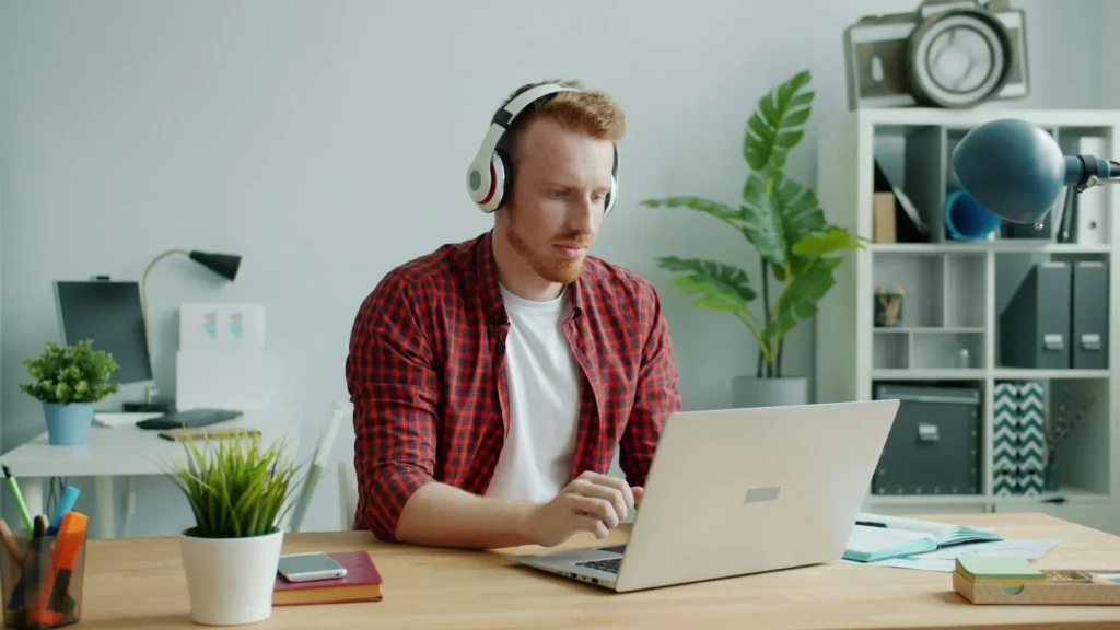 A person wearing headphones is sitting at a wooden desk, working on a laptop surrounded by plants and office supplies in a bright room.