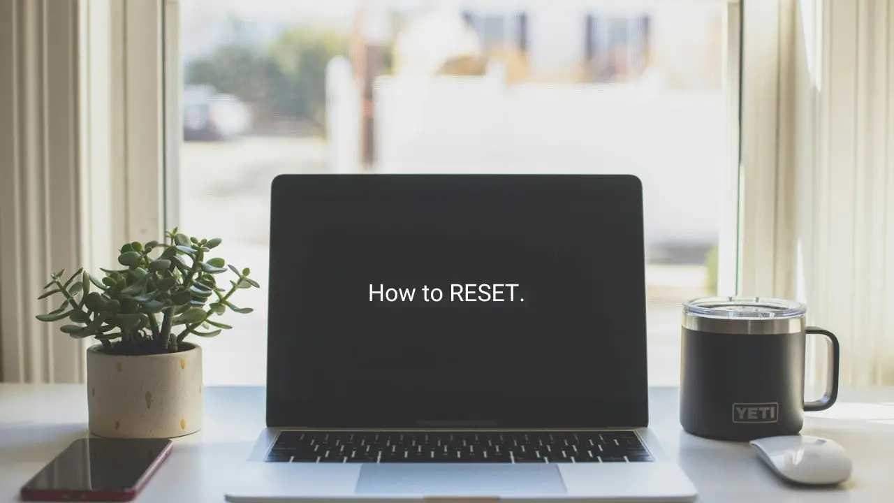A silver MacBook sits on a white desk with a potted plant, a smartphone, and a black YETI mug nearby. The laptop screen displays the text "How to RESET." in white letters against a black background, indicating a tutorial on resetting a device.