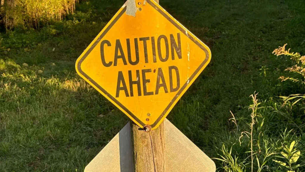 A worn yellow caution sign reading "CAUTION AHEAD" stands on a wooden post, surrounded by green grass and foliage.