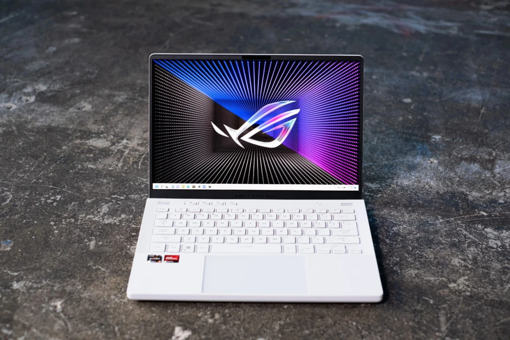 White gaming laptop placed on a dark concrete floor with the screen open showing a colorful radial light pattern and a stylized logo, white keyboard and large touchpad visible from the front