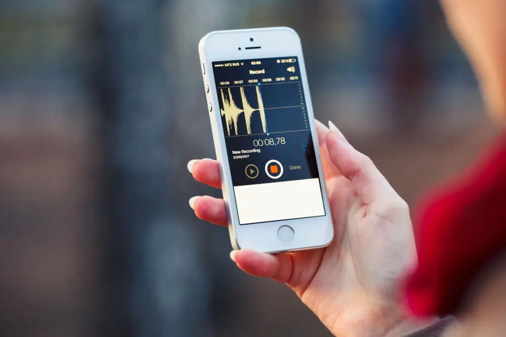 Hand holding an iPhone displaying the Voice Memos app with an active audio waveform recording on the screen