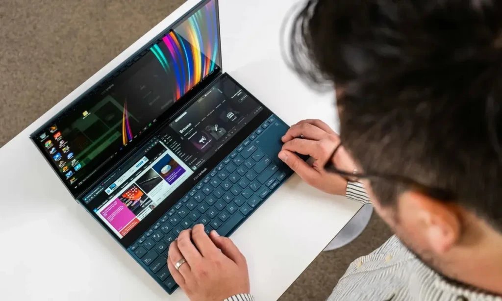Person seated at a white desk using a dual screen laptop with one screen above the keyboard, hands typing while multiple apps and windows are open across both displays, viewed from an overhead angle