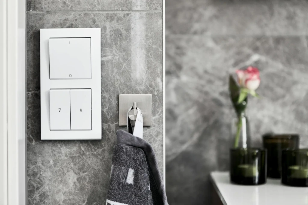 A modern light switch panel on a gray marble wall, with a towel hanging nearby and glass containers on a counter.