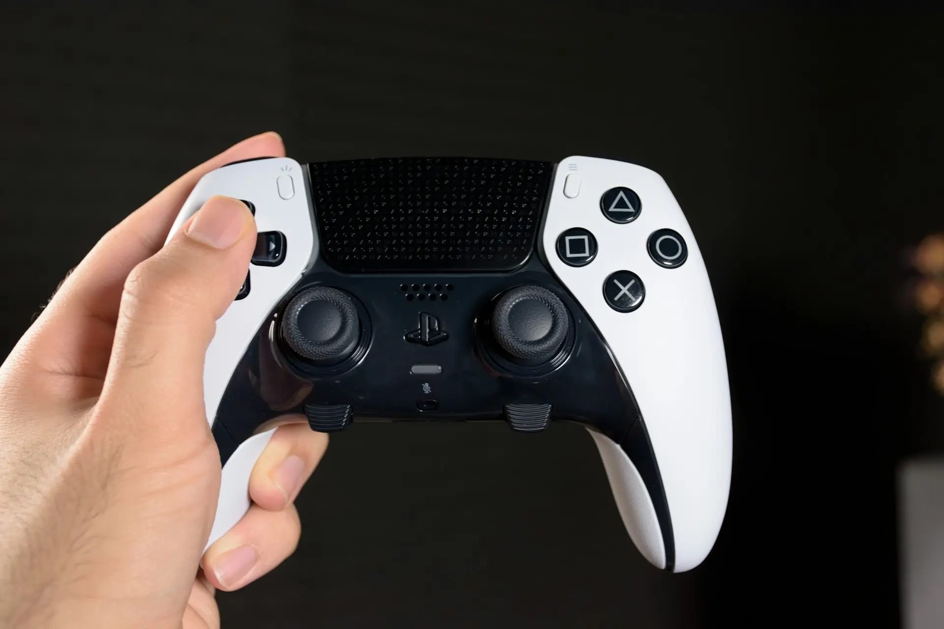 Close-up of a hand gripping a white PlayStation DualSense controller against a dark background, highlighting the textured grip, analog sticks, and iconic face buttons, emphasizing ergonomics and design for immersive gameplay