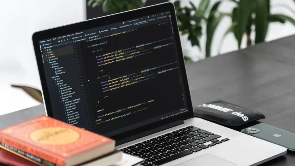 A silver laptop on a dark desk displaying JavaScript code in a dark-themed editor, surrounded by a stack of books, a smartphone, and a black pouch, suggesting a focused, real-world developer workspace blending learning and productivity.