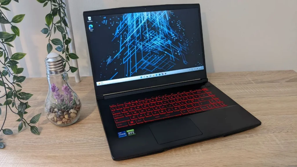 An MSI gaming laptop with red backlighting on its keyboard, running a blue circuit-themed wallpaper, set on a wooden desk beside a decorative lightbulb planter with green vines, showcasing its sleek design and aesthetic appeal in a modern home or office setup.