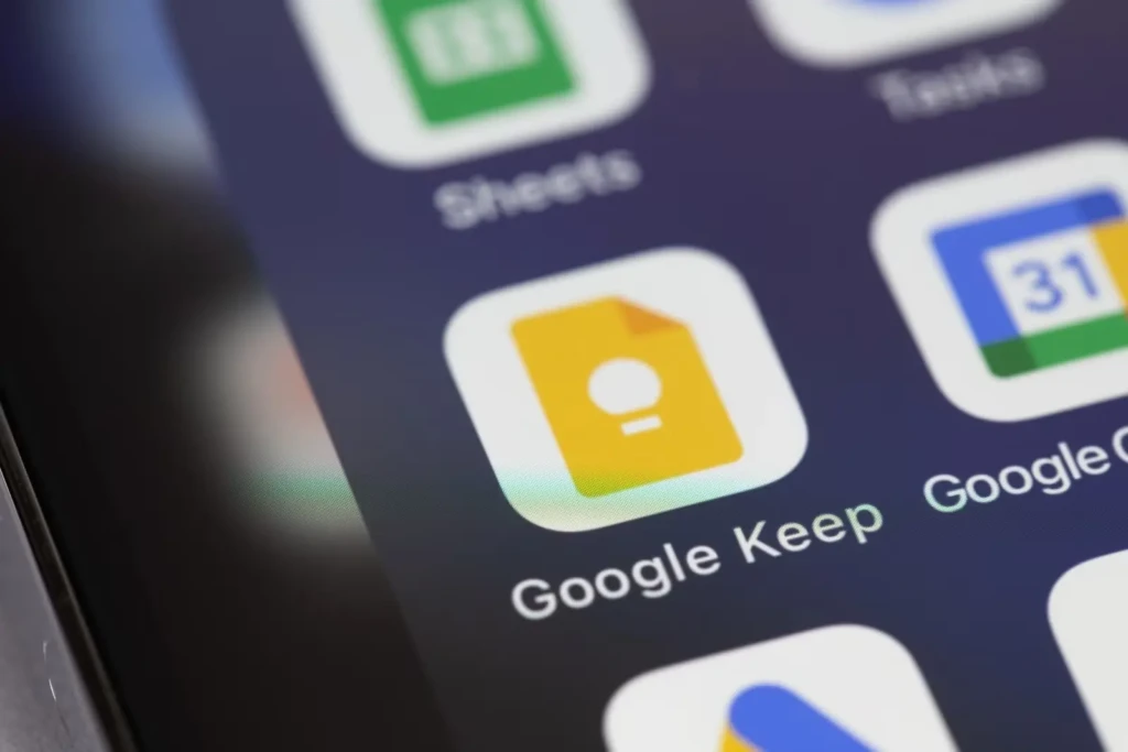 A close-up of a smartphone screen highlighting the Google Keep app icon, a yellow sticky note with a white lightbulb symbol, nestled among other Google apps like Sheets and Calendar. The focus emphasizes quick access to note-taking on mobile devices